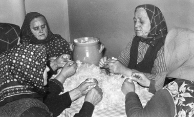 Idős asszonyok tollat fosztanak egy asztal körül ülve, hagyományos közösségi tevékenység.