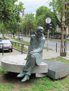 Arthur Koestler szobra egy kör alakú talapzaton ül, háttérben városi utca és fák.