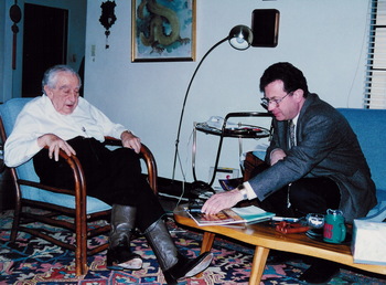 Két ember beszélget egy nappaliban, egyikük fotelben ül, másikuk kanapén, asztalon könyvek és bögre.