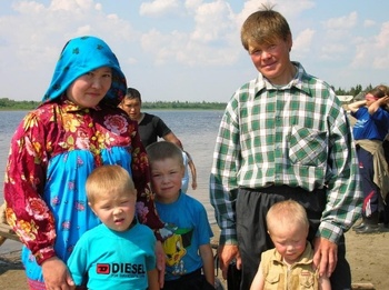 Egy család áll a tó partján, háttérben erdő és felhők láthatók. A gyerekek kék pólót viselnek, a felnőttek színes ruhákban vannak.