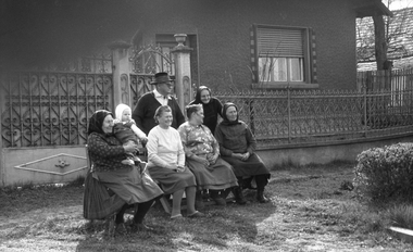 Család ül egy padon egy ház előtt, háttérben kovácsoltvas kerítés és ház látható.