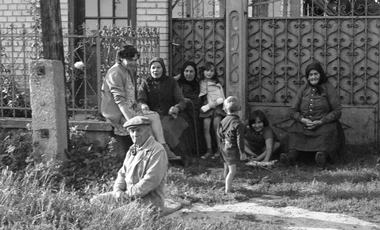 Csoport emberek ülnek és állnak egy kerítés előtt, gyerekek és felnőttek vegyesen.