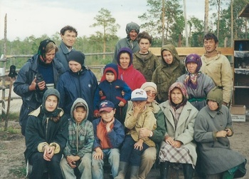 Csoportkép különböző korú emberekkel egy erdős területen, háttérben fák és kerítés.