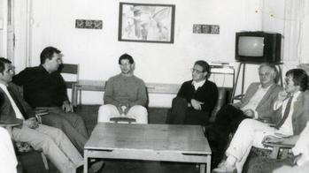 Csoportos beszélgetés egy szobában Csoport ül egy szobában, asztallal, háttérben televízióval és festménnyel.