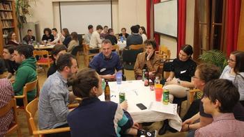 Fiatalok ülnek körbe asztaloknál egy beltéri helyszínen, az asztalokon italok és jegyzetek találhatók.