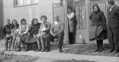 Csoportos kép, ahol emberek ülnek és állnak egy ház előtt, háttérben kerítés és épület.