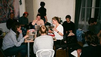 Csoportos megbeszélés laptopokkal egy asztal körül Csoport emberek egy asztal körül ülnek, laptopokkal dolgoznak vagy jegyzetelnek, háttérben festmény és táska.