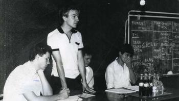 Csoport ül egy asztal körül, táblára írt szöveget néznek, üdítős üvegek az asztalon.