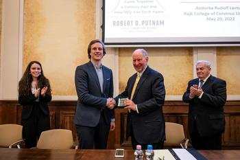 Díjátadó esemény Robert D. Putnam előadásán Két férfi kezet fog egy díjátadó eseményen, háttérben Robert D. Putnam előadásának címe és dátuma.