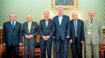 Hat elegánsan öltözött férfi áll egymás mellett egy díszes szobában, háttérben egy festmény.