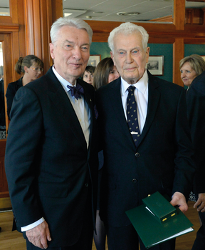 Két elegánsan öltözött férfi egy rendezvényen, egyikük kezében zöld mappa és doboz.