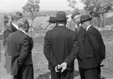 Öt elegáns öltözetű férfi áll egy kertben, háttal a kamerának, kalapban.