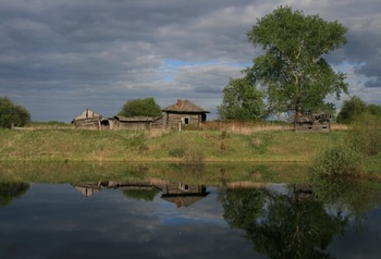 Elhagyatott vidéki ház egy tó partján, körülötte fák és bokrok, a víz tükrözi a tájat.