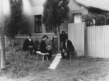 Csoport ember ül egy ház kertjében, körülvéve növényekkel és kerítéssel.