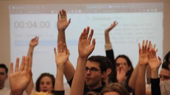 Emberek felemelik a kezüket egy előadás során Csoport ember felemeli a kezüket egy előadás vagy szavazás során, háttérben kivetítőn időzítő és szöveg.