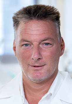 Fehér köpenyt viselő személy, akinek az arca el van homályosítva.