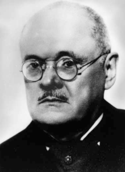 Fekete-fehér portré öltönyben és jelvénnyel Fekete-fehér portré, amelyen egy személy sötét öltönyt és jelvényt visel.