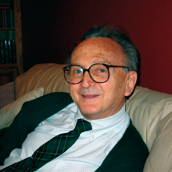 Férfi ül egy kanapén, fehér ingben és zöld kockás nyakkendőben, szemüveget visel.