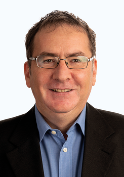 Férfi kék inget és fekete zakót visel, a képen a felsőtest látható.