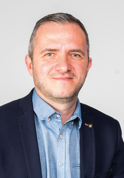 Férfi kék inget és sötétkék zakót visel, a képen a felsőtest látható.