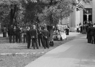 Férfiak öltönyben állnak egy templom előtt, háttérben Krisztus szobor és árusító stand.