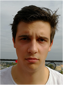 Fiatal férfi barna kócos hajjal, fehér pólóban, szabadtéri háttérrel