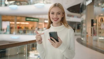 Fiatal nő okostelefont használ egy bevásárlóközpontban Fiatal nő fehér pulóverben, okostelefonnal a kezében, bevásárlóközpontban áll.