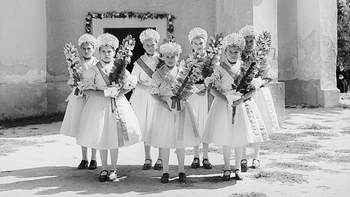 Fiatal lányok ünnepi ruhában virágokkal Hat fiatal lány ünnepi ruhában áll egy épület előtt, kezükben virágokkal.