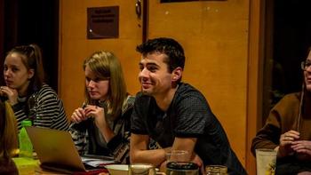 Fiatalok csoportja beszélgetés közben egy asztalnál Fiatalok csoportja ül egy asztal körül, laptop és italok az asztalon, háttérben felirat.