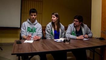 Három fiatal ül egy asztalnál, jegyzetfüzetekkel és tollakkal, Rajk feliratú pulóverben.