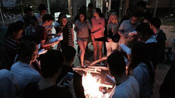 Fiatalok egy tábortűz körül állnak és énekelnek vagy beszélgetnek.