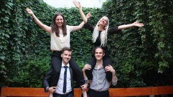 Fiatalok vállon ülve a szabadban Két fiatal férfi vállán ül két fiatal nő, háttérben zöld növények és egy pad.