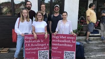 Fiatalok táblákkal, kérdésekkel és QR kódokkal Fiatalok táblákat tartanak, melyeken kérdések és QR kódok szerepelnek, háttérben egy ház.