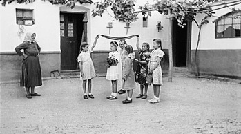 Gyerekek játék közben egy udvaron Gyerekek egy udvaron állnak, kendőt tartanak a fejük fölött, egy idős nő figyeli őket, háttérben egy ház.