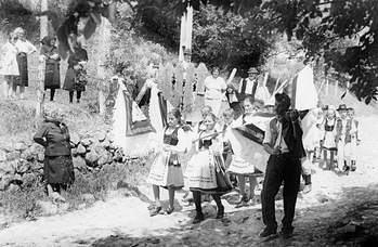 Hagyományos népviseletben vonuló csoport zászlókkal Hagyományos népviseletbe öltözött csoport zászlókkal vonul egy vidéki környezetben, nézők figyelik őket.