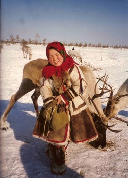 Hagyományos öltözetű személy rénszarvasokkal a havas tájon, háttérben fák és hó.