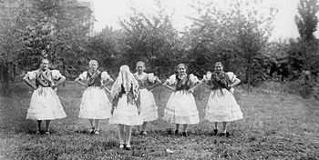 Fiatal lányok hagyományos népviseletben egy kertben Hat fiatal lány hagyományos népviseletben áll egy kertben, háttal a kamerának, körben elhelyezkedve.