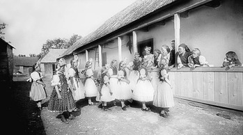 Hagyományos népviseletbe öltözött lányok egy parasztház előtt Hagyományos népviseletbe öltözött lányok sorban állnak egy parasztház előtt, vidéki környezetben.