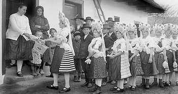 Hagyományos népviseletbe öltözött fiatalok ünnepi eseményen Fiatalok hagyományos népviseletben sorakoznak egy ház előtt, kosarat adva át ünnepi eseményen.