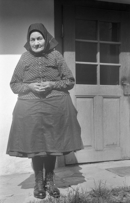 Egy hagyományos ruhába öltözött nő áll egy ajtó előtt, kendővel a fején és bakancsban.