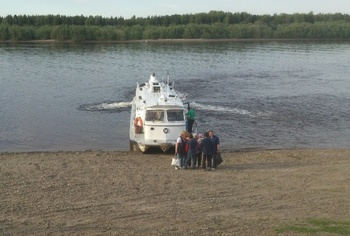 Hajó kikötése a folyó partján Hajó kikötött a folyó partján, emberek állnak a parton, háttérben fák és növényzet.