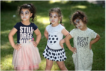 Három kislány feliratos pólókban áll a fűben Három kislány áll egymás mellett, különböző feliratú pólókban, a háttérben fű látható.