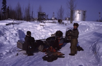 Három ember áll egy motoros szán mellett havas tájon, háttérben ipari létesítmények és fák láthatók.