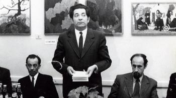 Hivatalos esemény beszéd közben, festményekkel a háttérben Férfi beszédet mond egy hivatalos eseményen, mellette két ülő férfi, háttérben festmények.