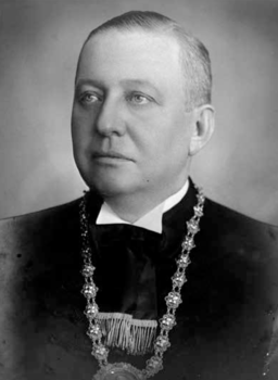 Hivatalos öltözetben lévő személy díszes lánccal Egy hivatalos öltözetben lévő személy, akinek a nyakában egy díszes lánc van.