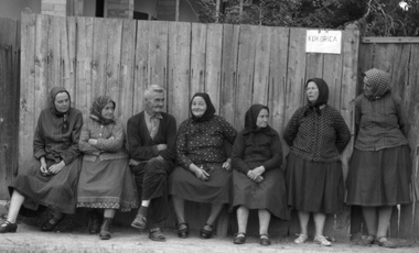 Idős emberek ülnek és állnak egy fa kerítés előtt, amelyen egy 'KUKORICA' feliratú tábla látható.