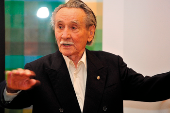 Idős férfi elegáns öltönyben gesztikulál Idős férfi elegáns öltönyben gesztikulál a kezével, háttérben színes fal.