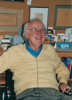 Idős férfi irodában képekkel és dokumentumokkal Idős férfi sárga pulóverben és kék ingben ül egy irodában, körülötte képek és dokumentumok.