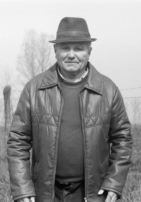 Idős férfi kalapot és bőrdzsekit visel, háttérben kerítés és fák láthatók.