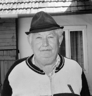 Idős férfi kalapot visel, és egy ház előtt áll, háttérben ablak és ajtó.